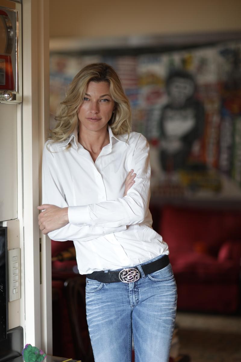 Nicole Junkerman in a white shirt and jeans, relaxed and leaning in a doorway beside colourful modern art and a red sofa at home.