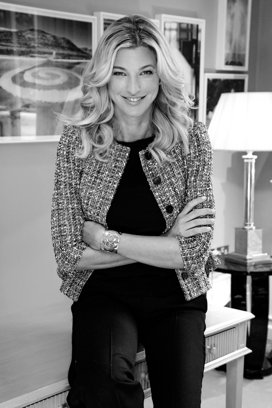 Nicole Junkerman in a tweed jacket over a black top, leaning against a table in a room hung with framed pictures, a black and white portrait.