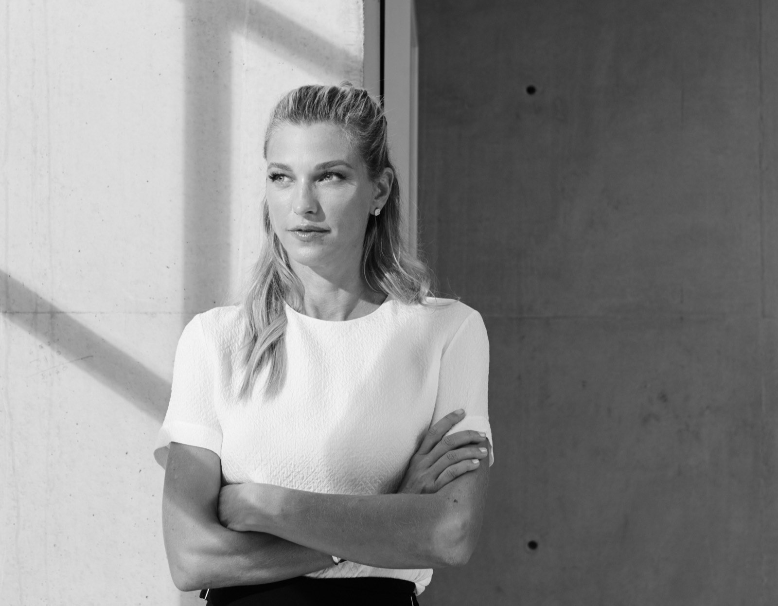 Nicole Junkerman in an ivory blazer with her arms crossed, a sleek editorial portrait against a concrete wall.