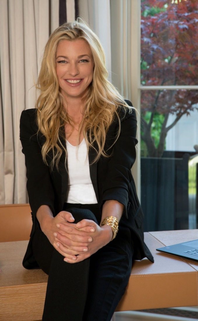 Nicole Junkerman smiling, seated on a desk by a window in a black blazer and white top.