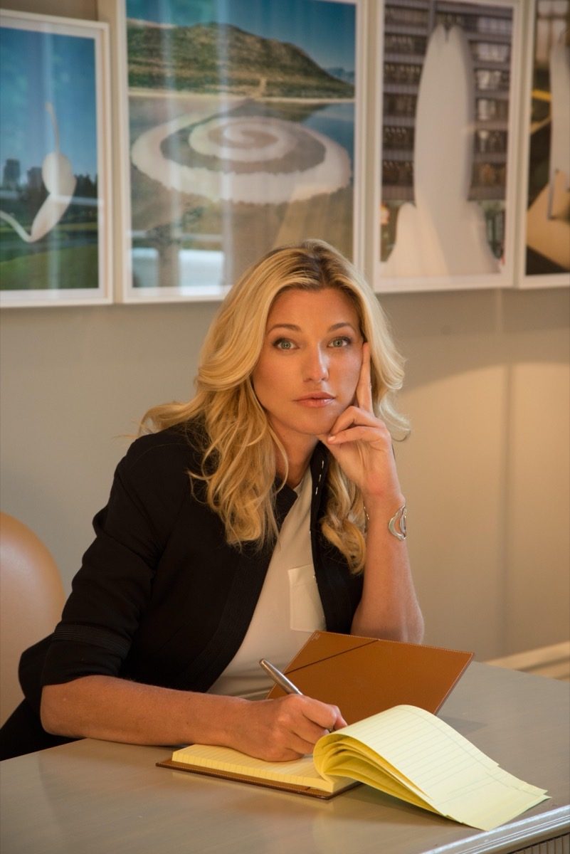 Nicole Junkerman in a black blazer writing thoughtfully in a notebook at her desk, with framed prints on the wall behind.