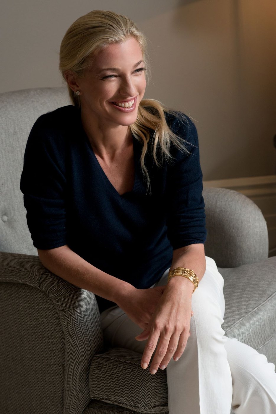 Nicole Junkerman seated in a pale armchair in a navy jumper, laughing and looking off to one side in soft indoor light.